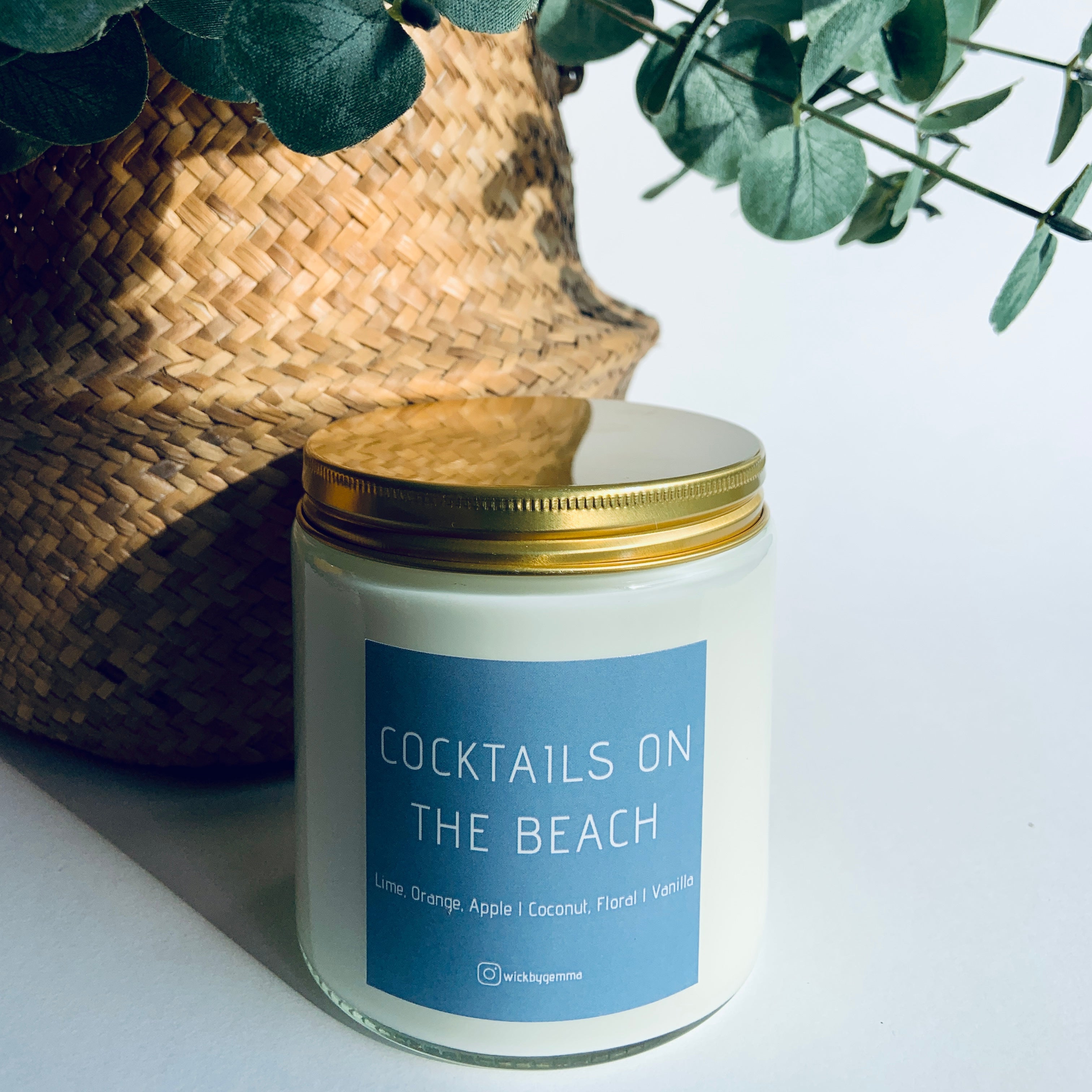 Candle labeled 'Cocktails on the Beach' with a woven basket and eucalyptus leaves in the background.