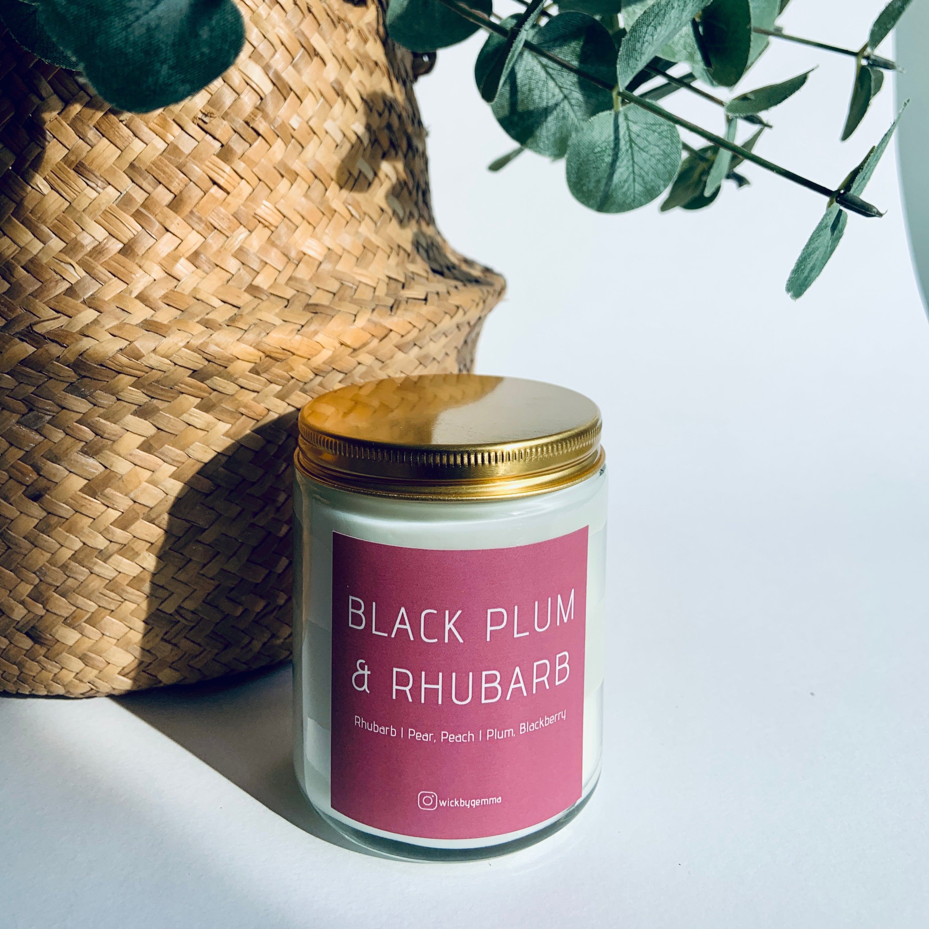 Candle labeled 'Black Plum & Rhubarb' with a gold lid next to a woven basket and green leaves on a light background