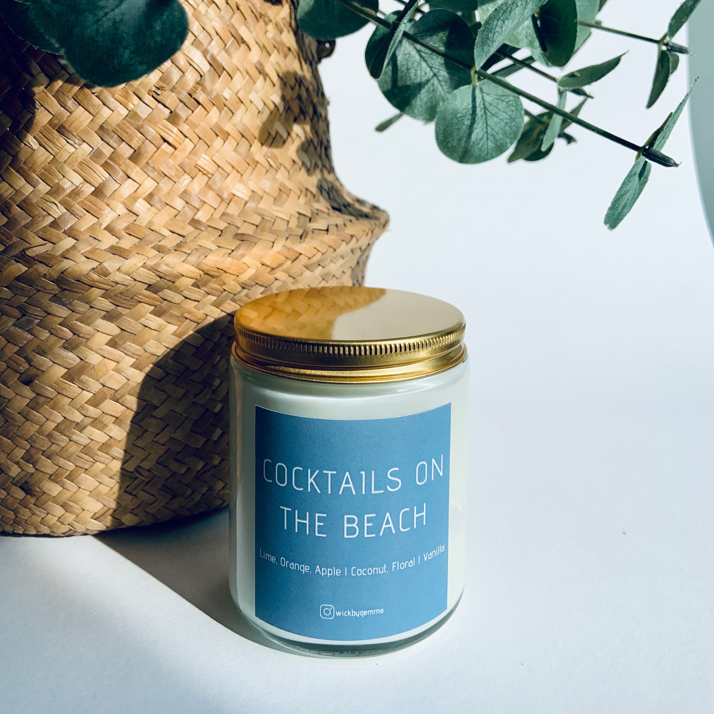 Candle labeled 'Cocktails on the Beach' next to a woven basket with greenery