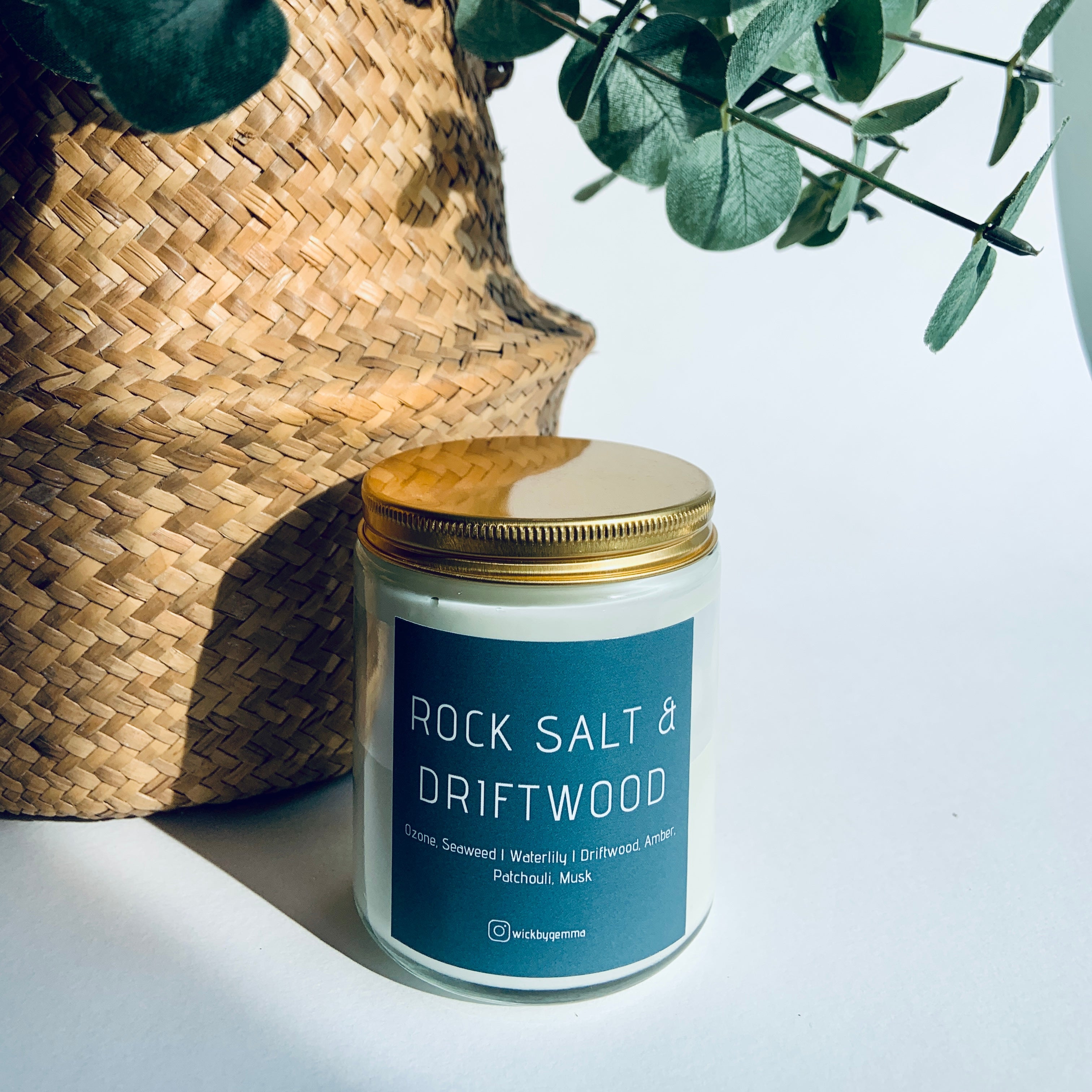Candle labeled 'Rock Salt & Driftwood' with a woven basket and eucalyptus leaves on a light background