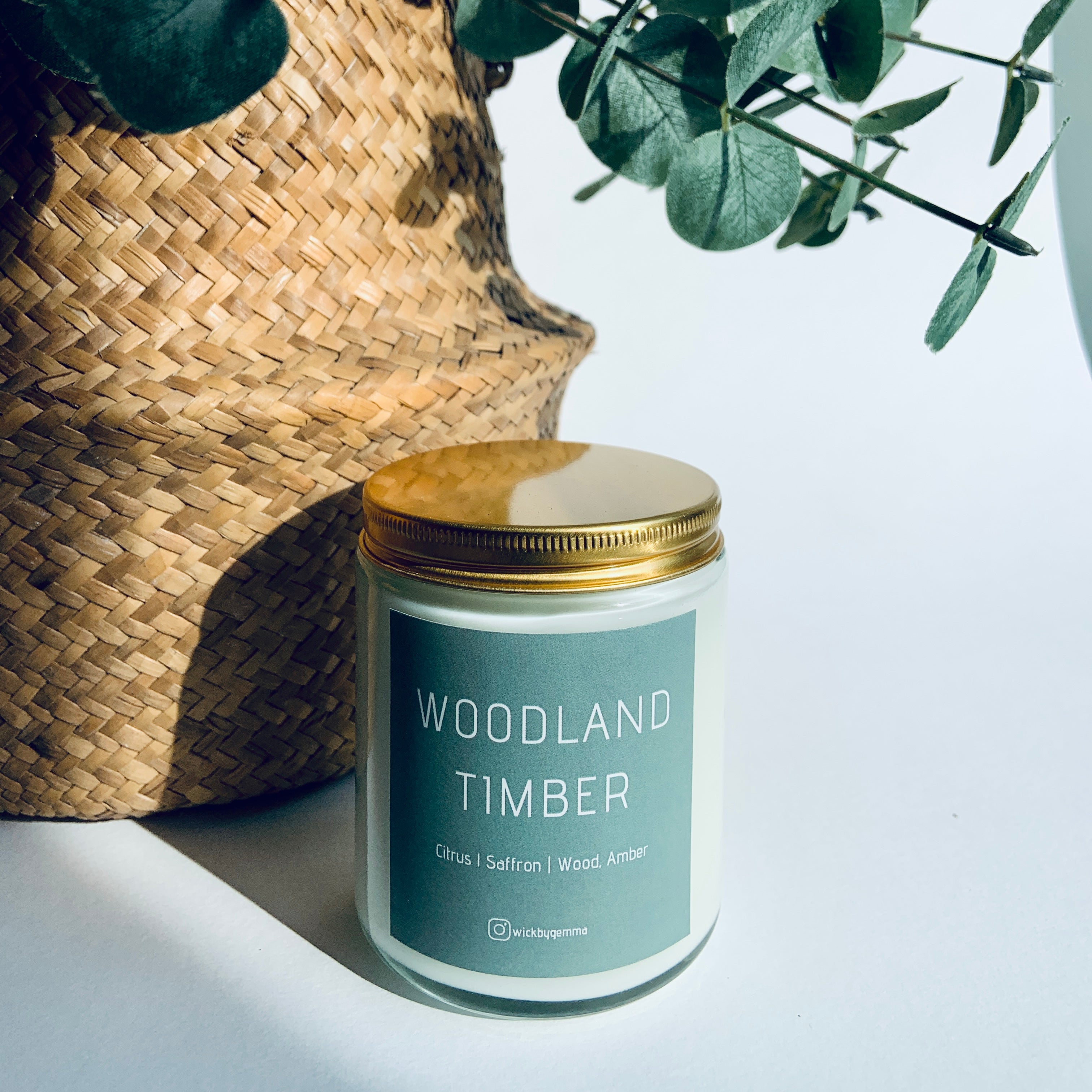 Candle labeled 'Woodland Timber' next to a woven basket with greenery on a light background