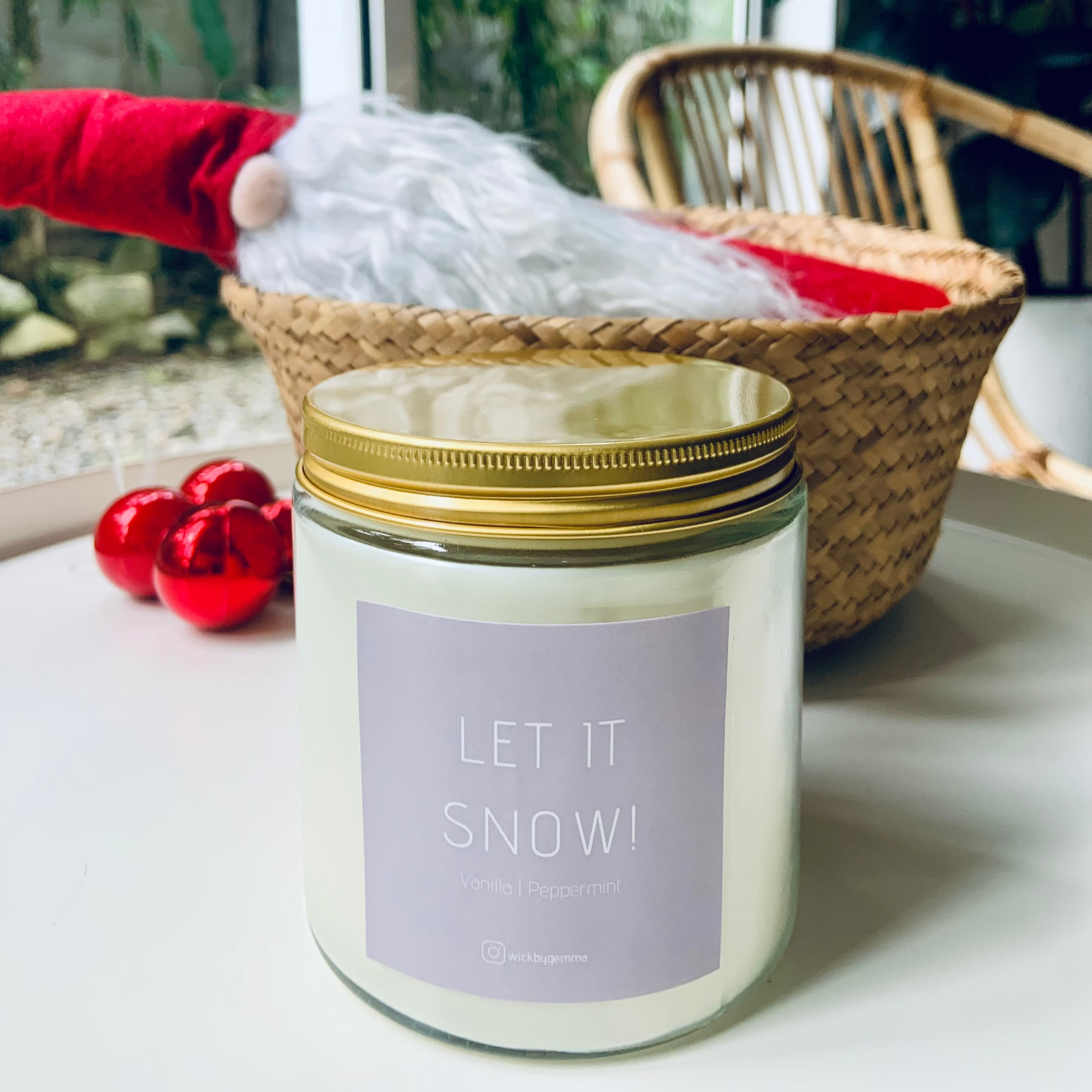 Candle labeled 'Let It Snow' with a gold lid on a white surface, with a blurred background of a window and plant.