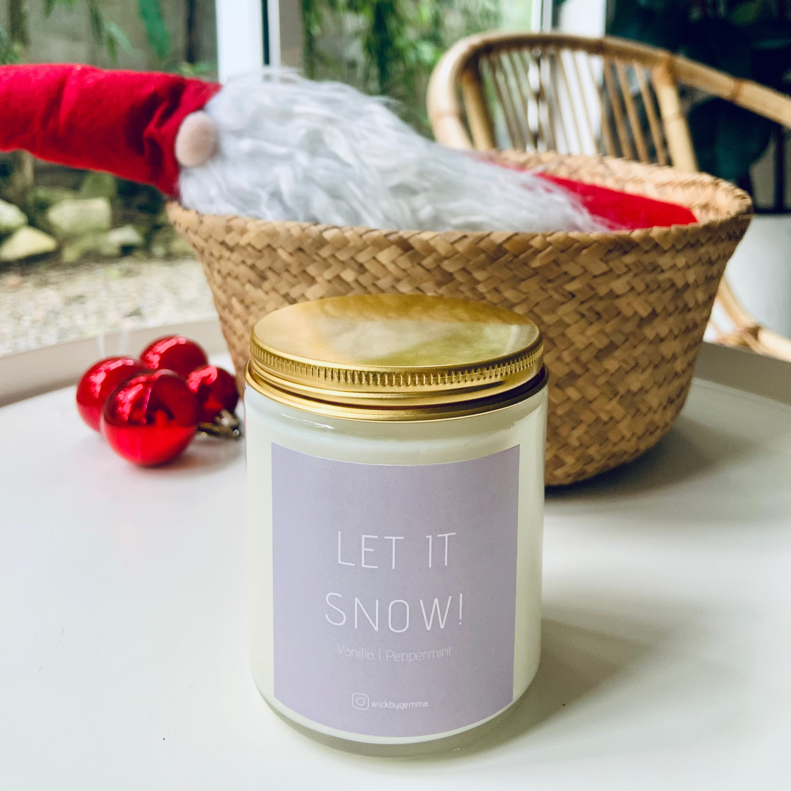 Candle with 'Let It Snow!' label on a surface near a window with plants