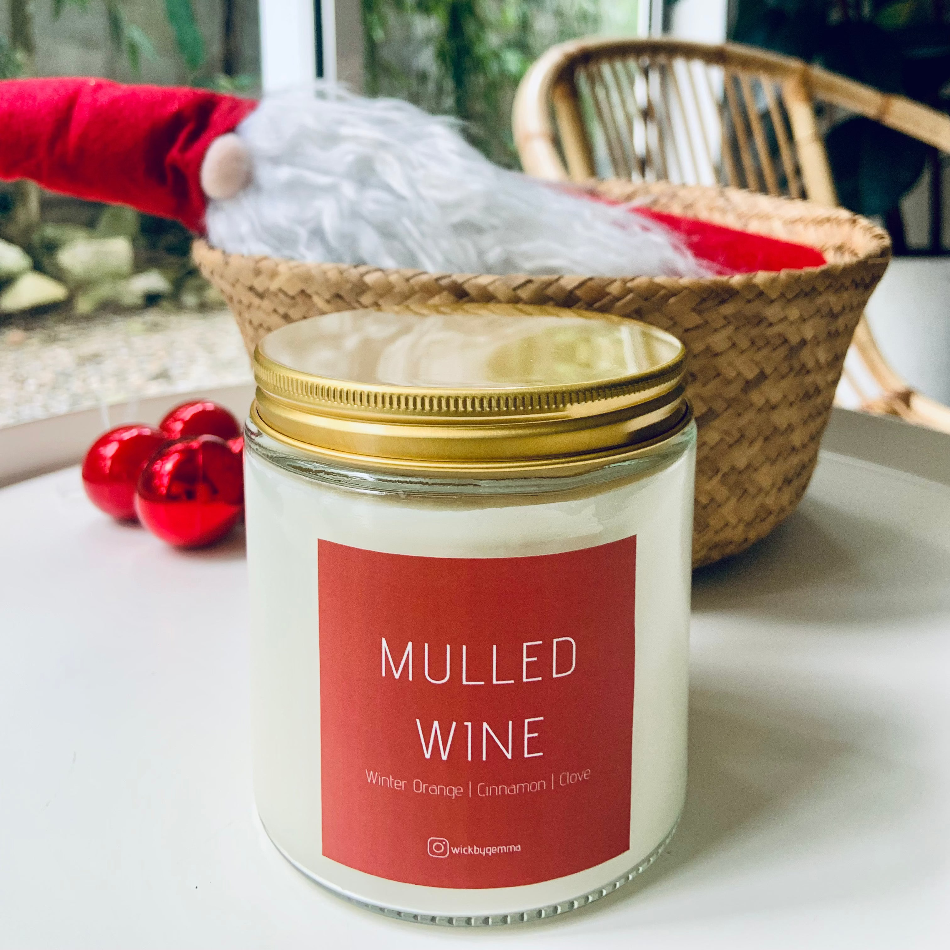 Candle labeled 'Mulled Wine' with a red label on a white surface, with a blurred background of a window and plant.