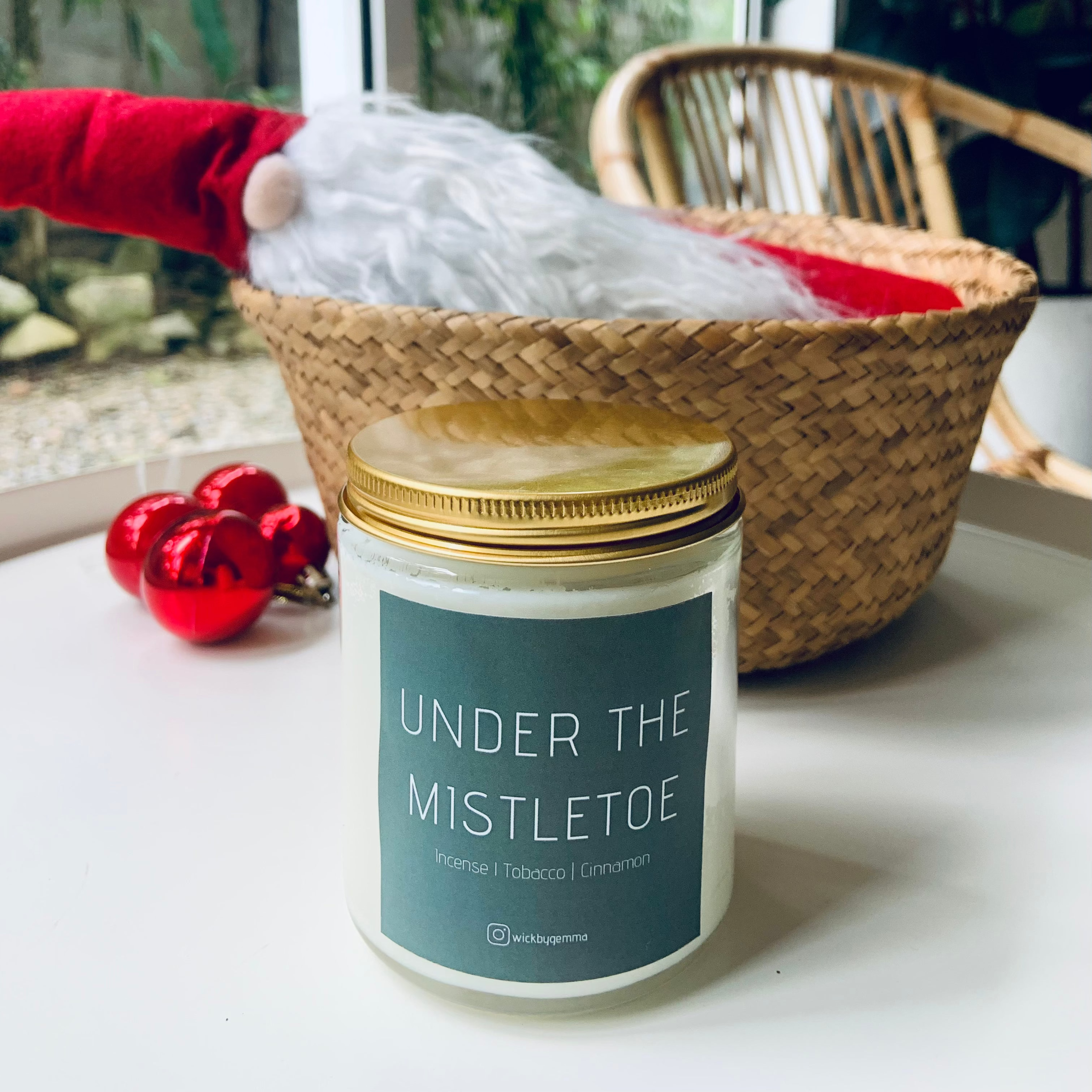Candle labeled 'Under the Mistletoe' on a surface with a decorative basket and red ornaments in the background.